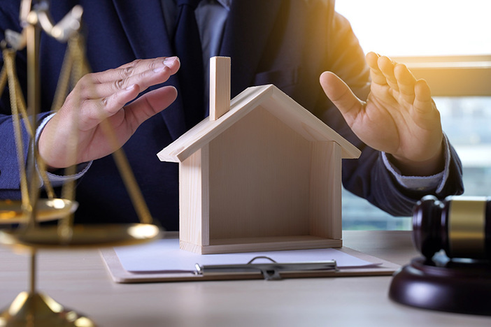 4 Different Types of LLCs and the Ways They Pay Taxes man in suit sitting at desk sheltering wooden model home on desk with hands, gavel and scale of justice in view