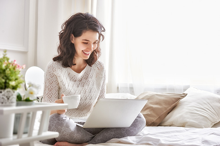 4 Simple Steps Anyone Can Use to Purchase Rental Properties Happy casual beautiful woman working on a laptop sitting on the bed in the house.
