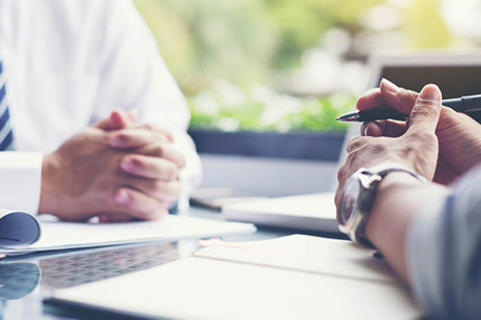 The Biggest Advantage of Doing Due Diligence Well close up of two men sitting on either side of desk with hands on desktop clasped one holding pen with hands resting on binder