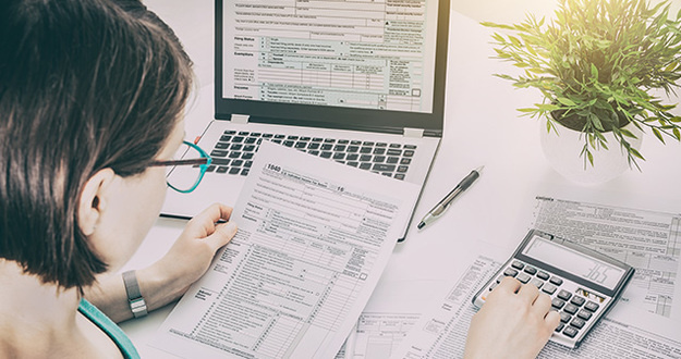 Multifamily Investors: Here’s Why Cost Segregation is Your Friend woman at desk with paper tax forms, tax form on lap screen, calculator, pen, plant
