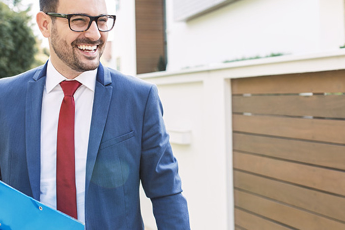 4 Simple Steps to Assess Reality & Make an Offer in a Seller’s Market smiling brown-haired man with glasses and shadow of a beard evaluates exterior of a residential property while wearing suit and carrying a folder