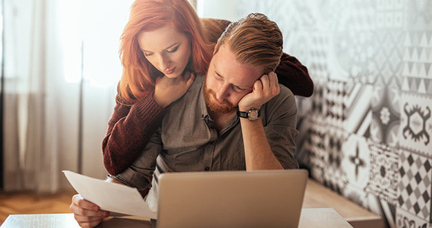 The 12 Most Common Financial Mistakes Made by Couples—and How to Avoid Them tired, frustrated young couple going over bill man sitting at desk with laptop woman looking over his shoulder
