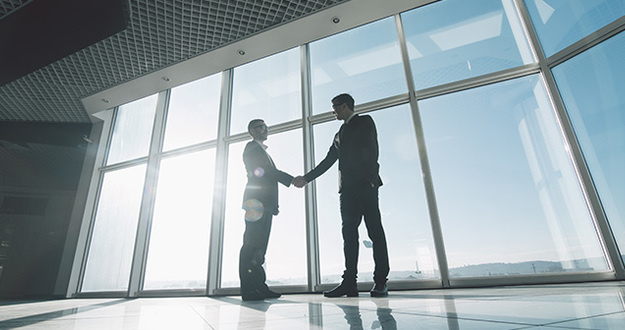 Should You Invest in Commercial Properties? 10 Investors Weigh In Two young businessmen are shaking hands with each other standing against panoramic windows.