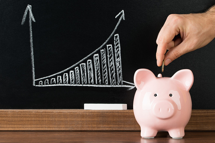 Traditional 401(k) vs. Roth IRA: Which One Wins? Hand Putting A Coin Into Piggy Bank In Front Of Blackboard Showing Graph