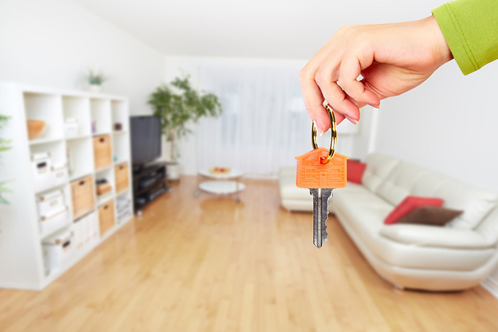 7 Ways to Buy Real Estate With Little (or No) Money Hand with house key in foreground, living room with light wood floors, white couch and white shelves in background