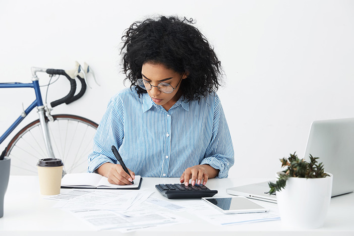 Introduction to Real Estate Investment Analysis Portrait of attractive hardworking businesswoman with Afro hairstyle busy doing paperwork at office desk, working through finances, using calculator and making notes in her notebook with pen