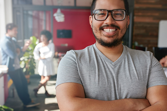 3 Steps to Close Your First Wholesaling Deal (& Find a Mentor in the Process) Smiling male entrepreneur looking at camera at office. Mixed race male creative professional with colleagues talking in background.